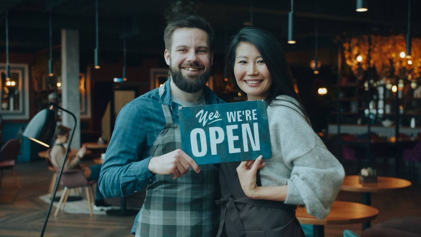 Smiling cafe owners holding an open sign inside their cozy cafe, welcoming customers warmly.