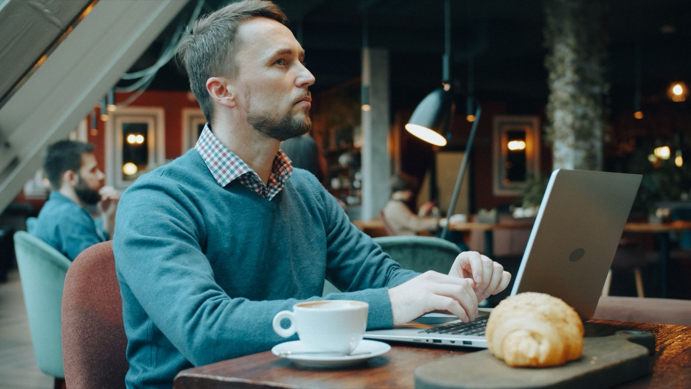 A man in a cafe working on a laptop with a coffee and croissant on the table.