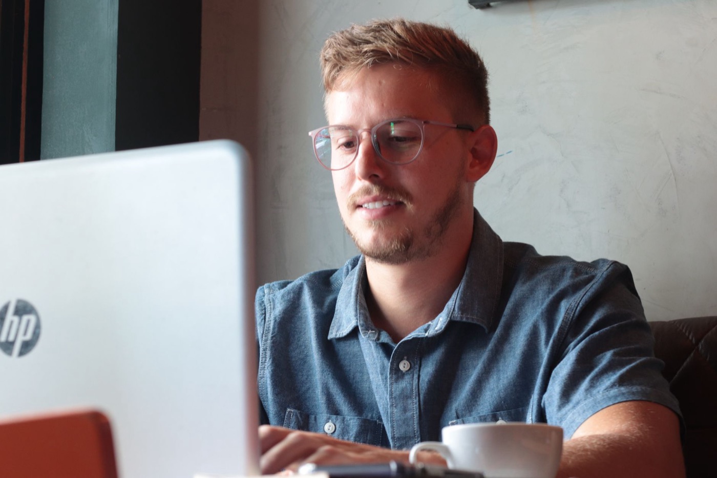 Male remote employee in eyeglasses surfing internet on netbook while sitting at table with cup of hot drink in cafeteria.